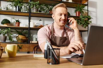A man busily works at his laptop in a plant shop while having a phone conversation.