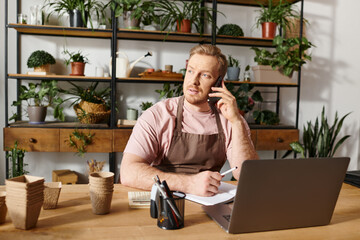 A man sits at a table in a plant shop, talking on a cell phone as he manages his small business.