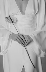 Naklejka premium A close-up, black-and-white photograph of a person wearing an elegant white dress with a deep V-neckline and long, flowing sleeves. The individual is holding a single anthurium flower