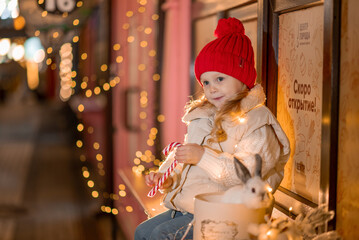 A little girl, wearing a bright red hat, beams with  a fluffy white rabbit, surrounded by the festive bustle of a Christmas market.