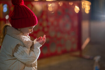 A little girl, wearing red hat,  blows playfully on her palm, sending sparkling snowflakes scattering through the air.