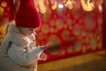 A little girl, wearing red hat,  blows playfully on her palm, sending sparkling snowflakes scattering through the air.