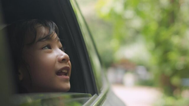 Joy of a road trip of  adorable Asian girl peering out a car window. Set against lush greenery, it shows adventure, curiosity, and childhood innocence on travel, family, nature, and happy moments.