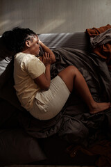 Top view of young depressed woman with legs bent in knee and eyes closed lying on bed and suffering...