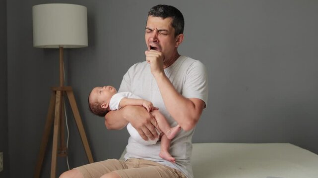 Young sleepy exhausted father holding cute little newborn baby in his arms with care man yawning covering mouth with palm sitting on bed in home interior