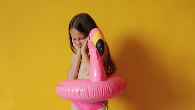 Bored sad little girl in flamingo rubber ring around waist isolated over yellow background feeling lonely has no friends to play with touching her cheeks expressing sadness and sorrow