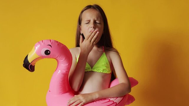 Sleepy tired brown haired little girl in flamingo rubber ring around waist isolated over yellow background standing covering mouth while yawning needs rest