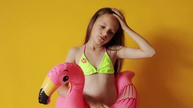 Sad tired sick little girl in flamingo rubber ring around waist isolated over yellow background suffering headache after long hours under son having sunstroke