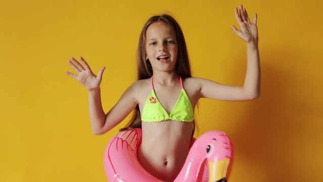 Cheerful little girl in flamingo rubber ring around waist isolated over yellow background celebrating her summer vacation resting in resort on sea shore dancing rejoicing