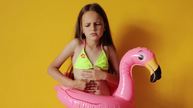 Ill sick little girl in flamingo rubber ring around waist isolated over yellow background suffering terrible stomach ache having food poisoning by seafood
