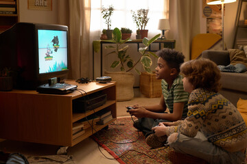 Two multiethnic boys engaged in playing a retro video game on an old television set, sitting on a...