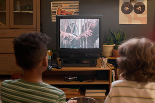 Back view of two kids sitting on floor watching TV showing horror movie about zombies in cozy retro living room with plants and posters on wall, and old-fashioned gaming console below TV screen