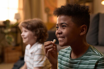 Two multiethnic boys smiling and watching TV while sitting on floor in cozy living room, enjoying time together with snacks and a relaxed atmosphere