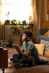 Vertical shot of African American boy sitting on retro carpeted floor playing arcade 8bit video...