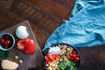 Fresh pasta salad bowl on a wooden background. Spinach, tomato, onion bell pepper, gouda, cheese, and multicolored, spiral pasta in a dark blue bowl.
