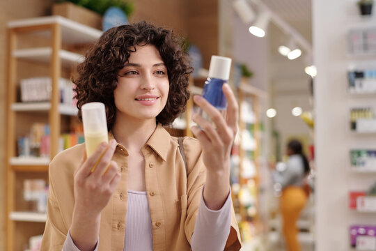 Happy young brunette woman in casualwear choosing makeup remover while standing in cosmetic supermarket and looking at bottle