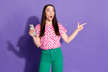 Photo portrait of pretty young girl hold device point look empty space wear trendy pink outfit isolated on purple color background