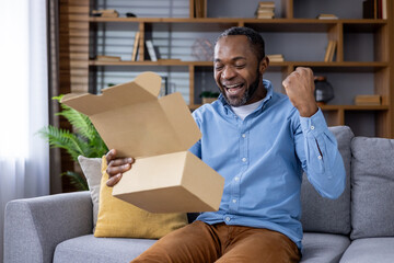 Man joyfully opening cardboard box while sitting on couch in modern living room. Displaying excitement, happiness, and surprise. Concept of receiving gift, positive news, or unexpected delivery.