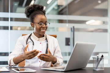 Professional doctor sitting at desk conducting video call consultation in modern office. Stethoscope around neck, gestures to communicate effectively. Medical professional using technology