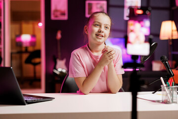 Joyful small girl shooting video for social media platform using smartphone. Young media star entertaining followers, doing content creation in pink neon lit apartment using phone on tripod