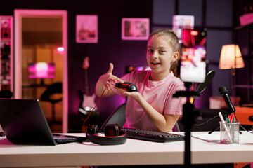 Cute girl in studio reviewing neon lit gaming mouse in front of camera in personal studio. Joyous kid filming electronics haul, presenting Bluetooth wireless computer peripheral to viewers