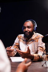 African American man creates internet radio programs at home with audio gadgets. Black male content creator using wireless headphones, a microphone, and podcast equipment while talking with guest.