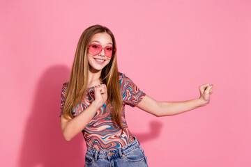 Portrait of adorable funky teen with long hairstyle wear colorful shirt in heart shape glasses dancing isolated on pink color background
