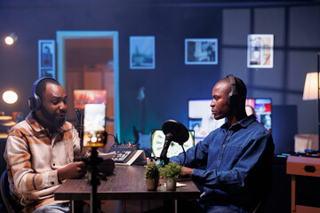 Vlogger is recording an interview with smartphone on tripod in studio while reviewing notes to ask quality questions to guest. Journalist using mobile device to film vlog while talking on mic with man