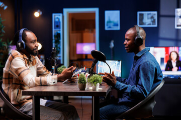 Two african american men are having serious conversation and taping it for online audience. Black male content makers sit at home and use audio equipment to produce a live radio show.