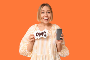 Surprised adult woman holding speech bubble with word OMG and mobile phone on orange background