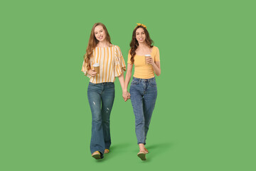 Beautiful young sisters with cups of coffee walking on green background