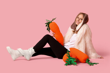 Beautiful woman in bunny ears with toy carrots sitting against pink background. Easter celebration