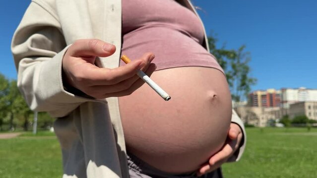 Close up of big belly of young pregnant woman is smoking cigarette in summer park