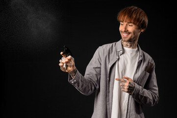 Young tattooed barber pointing at hair spray on dark background