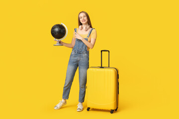 Smiling young woman with headphones pointing at globe on yellow background. Travel concept