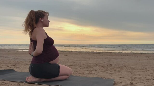Young pregnant active woman doing exersice outdoors, stratching her body and meditate on the sea, beach at sunset in the evening
