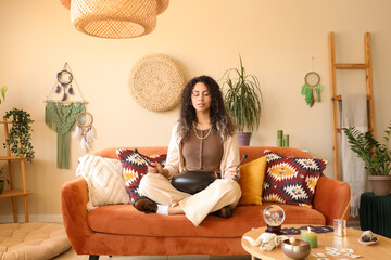 Young African-American witch with glucophone meditating on sofa at home