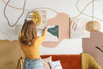 Young woman hanging stylish painting on wall at home, back view