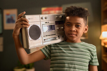 Portrait of African American boy holding boombox on shoulder in living room, looking at camera with slight smile