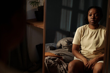 Mirror and reflection of young desperate African American woman sitting on bed and looking at...