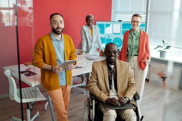 Group of diverse colleagues posing in modern office with presentation screen in background. Black man in wheelchair sits at front, while others stand around table, smiling