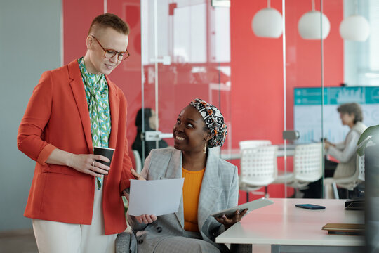 Caucasian female and African American female colleagues discussing work while reviewing documents in office, one holding tablet and other holding coffee cup