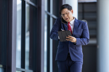 Fototapeta premium Middle-aged Asian businessman rushing with work on his way to office. Male HR officer uses tablet and hurry to find information about employees in corporate. Man manager working on tablet.