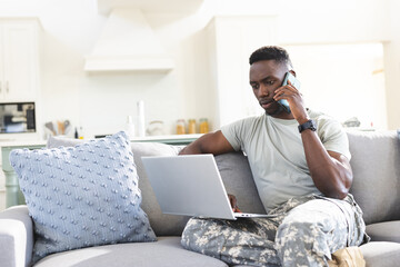 African american male soldier on couch at home using laptop and talking on smartphone, copy space