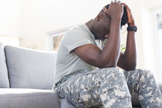 Troubled african american male soldier sitting on couch at home, holding head, copy space