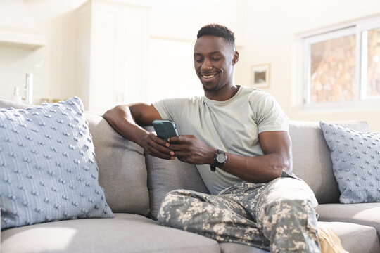 Happy african american male soldier sitting on couch at home using smartphone and smiling