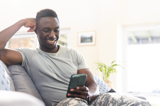 Happy african american male soldier sitting on couch at home using smartphone, smiling, copy space