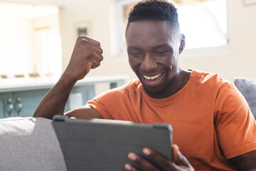 Happy african american man sitting on sofa watching tablet and celebrating in sunny living room