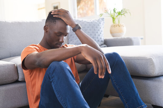 Thoughtful african american man sitting on floor holding head in sunny living room - Powered by Adobe