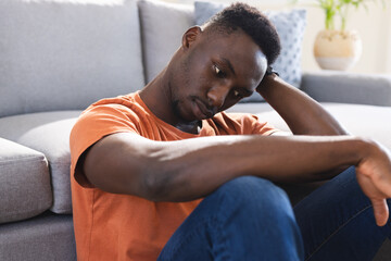Thoughtful african american man sitting on floor in sunny living room holding head